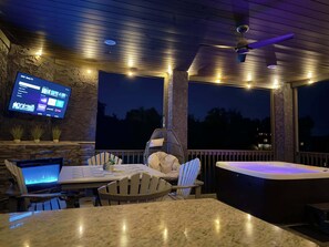 A covered outdoor area with a stone wall, a flat-screen TV mounted on the wall, a hot tub, and a table with chairs. - Lux Treehouse Cabin, Hot Tub, Sauna, Arcade (Sevierville)