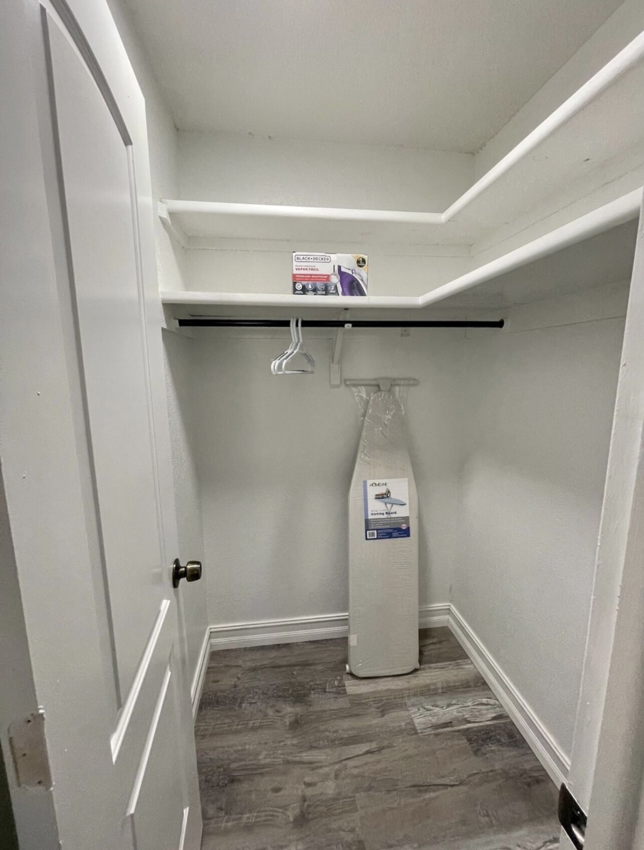 A small, narrow room with white walls and a wooden floor. There is a door on the left, a shelf with a box on it, and a bottle of cleaning product on the floor.