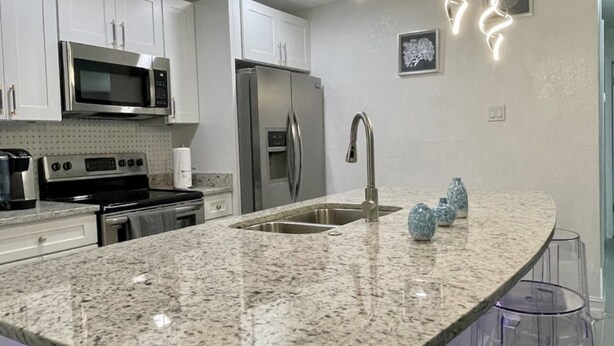 A modern kitchen with white cabinets, stainless steel appliances, a granite countertop, and pendant lights. - Relaxing Home Near Universal (Orlando)