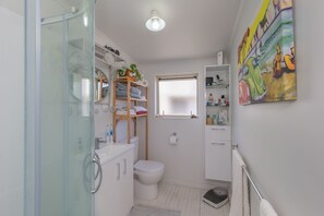 Un baño pequeño con cabina de ducha de vidrio, inodoro blanco, una ventana discreta y un estante con artículos de aseo.