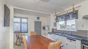 Een keuken met een houten eettafel, rieten stoelen, witte kasten, een hangende pannendroger en uitzicht door het raam.