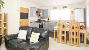 A compact living space with a black leather sofa, a wooden dining table with chairs, a kitchen area with grey cabinets, and a white refrigerator. - 3Bed/2Bath Duplex Apartment - Pure Bliss Belfast (Belfast)