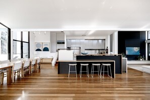 A modern kitchen with a central island, bar stools, and a dining area with a long table and chairs. - Luxury & Breathtaking ocean views (Sunshine Beach)