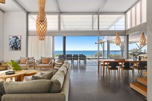 A modern living room with a large sofa, dining table, and chairs, leading to a balcony with a view of the sea.