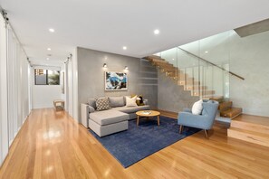 Uma sala de estar moderna com um sofá cinza, uma poltrona azul e uma mesa de centro de madeira. Uma escadaria com guarda-corpo de vidro dá acesso a um andar superior.