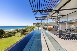 A modern infinity pool with a clear view of the ocean, a covered outdoor dining area, and a contemporary kitchen visible through large glass doors.
