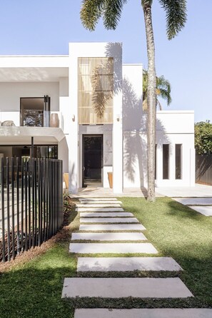 A modern house with a concrete pathway leading to the entrance, surrounded by well-maintained grass and palm trees.