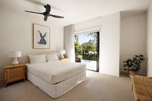 A hotel room with a large bed, a ceiling fan, a framed artwork, a nightstand, a lamp, a view of the outdoors through a sliding door, and a potted plant.
