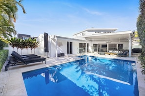 A modern outdoor pool area with lounge chairs, a covered seating area, and a large house in the background.