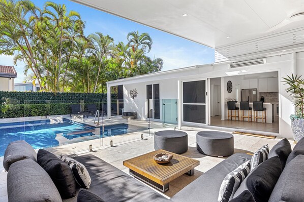 A modern outdoor living area with a pool, seating, and a kitchen visible through an open door.