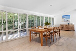 A dining area with a wooden table and chairs, large windows with blinds, and a painting on the wall.