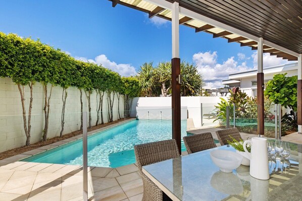 A pool area with a table and chairs, surrounded by a white wall and greenery.