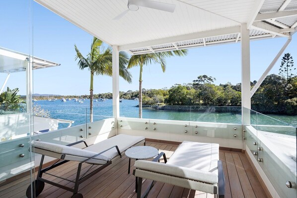 A sunlit deck with lounge chairs and a small table, overlooking a marina with boats and palm trees.