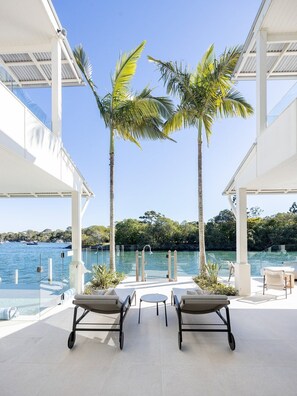 An outdoor seating area with two lounge chairs and a small table, overlooking a body of water with boats and greenery in the background. - Luxurious Riverfront Retreat (Noosa Heads)