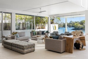 A modern living room with large windows, a ceiling fan, and a view of the sea. - Luxurious Riverfront Retreat (Noosa Heads)