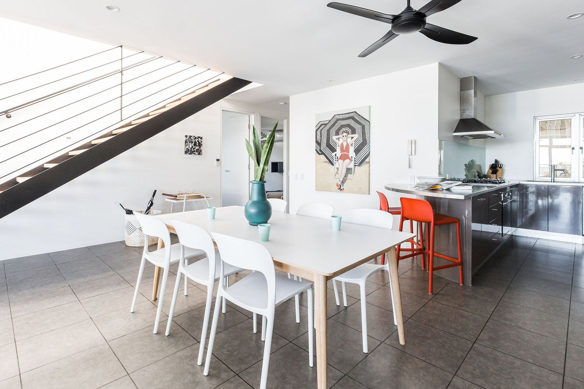 A modern kitchen with a central island, a dining area with white chairs, and a staircase with a glass railing.