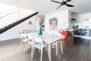 Une cuisine moderne avec un îlot central, un coin repas avec des chaises blanches et un escalier doté d’une rampe en verre.