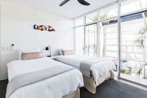 A hotel room with two beds, a ceiling fan, a wall-mounted artwork, and a large window with a view of palm trees.