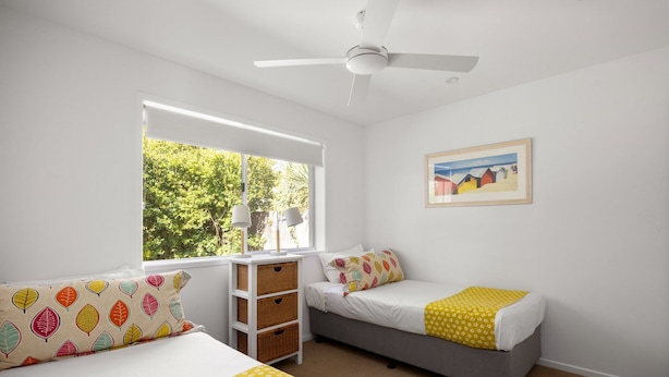 Ein Schlafzimmer mit zwei Betten, einem Deckenventilator, einem Fenster mit Blick auf Bäume und einem gerahmten Bild an der Wand.