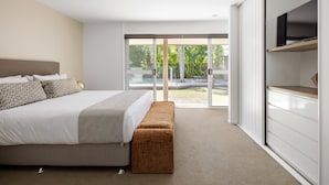 Ein modernes Schlafzimmer mit einem großen Bett, einer Bank, einem Fernseher und einer Glastür, die auf einen Balkon mit Blick auf Bäume und einen Pool führt.