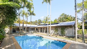 Ein Pool mit Holzterrasse, umgeben von Palmen und einem Gebäude mit großen Fenstern.