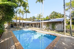 A swimming pool with a wooden deck, surrounded by palm trees and a building with large windows.