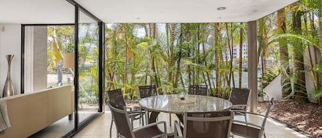 Una mesa de vidrio redonda con sillas en una habitación con ventanas amplias que dan a un jardín tropical.