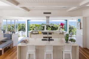 A modern kitchen with a large island, white bar stools, and a view of the ocean. - The Beach Shack - Getaway on Sunshine Beach (Sunshine Beach)