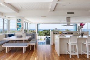 A modern kitchen with a view of the sea, wooden flooring, and a dining area with striped cushions. - The Beach Shack - Getaway on Sunshine Beach (Sunshine Beach)