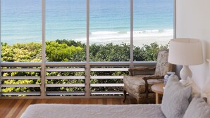 Ein Zimmer mit großem Fenster mit Blick auf einen Strand, ein Sessel, eine Lampe und ein weißes Sofa mit Struktur.