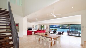 Ampla sala de estar com grande mesa de jantar, sofá vermelho e vista para a água por grandes janelas.
