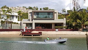 Uma casa moderna de dois andares com varanda, um barco na água e uma praia ao fundo.