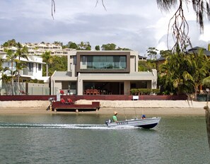 A modern two-story house with a balcony, a boat on the water, and a beach in the background.