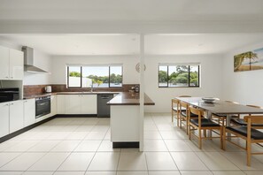 A modern kitchen with white cabinets, a central island, and a dining area with wooden chairs. - Breathtaking River Views on Noosa Parade (Noosa Heads)