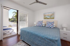 A bedroom with a bed, two pillows, a nightstand, a lamp, a ceiling fan, a window with blinds, and a painting on the wall. - Breathtaking River Views on Noosa Parade (Noosa Heads)