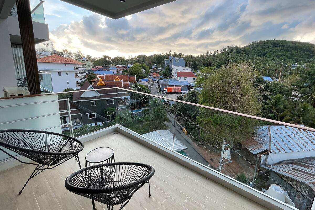 Un balcon donnant sur la ville, avec une balustrade en verre, deux chaises noires en fil de fer et une petite table.