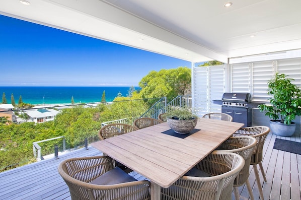 A deck with a wooden table and wicker chairs, overlooking a beach and ocean.