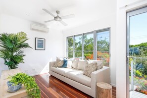 A modern living room with a sofa, a ceiling fan, a large window, and a potted plant.