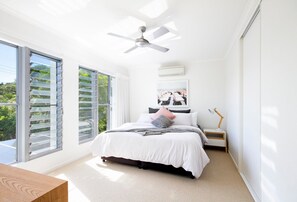 A bedroom with a bed, a ceiling fan, a nightstand, a lamp, and a large window with blinds.