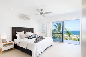 Une chambre avec un lit, une table de chevet, une lampe, un ventilateur de plafond et une vue sur l’océan depuis un balcon.