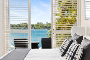 A bedroom with a view of a beach and boats, a bed with a gray blanket, and a black chair. - Luxury Riverfront: Private Pool, Water Views (Noosa Heads)