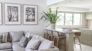 A modern living room with a sofa, a coffee table, and a dining area with bar stools. There are framed pictures on the wall and a potted plant on the table. - Luxury Riverfront: Private Pool, Water Views (Noosa Heads)