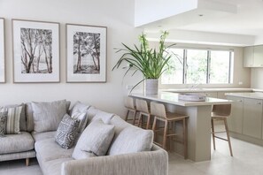 A modern living room with a sofa, a coffee table, and a dining area with bar stools. There are framed pictures on the wall and a potted plant on the table. - Luxury Riverfront: Private Pool, Water Views (Noosa Heads)