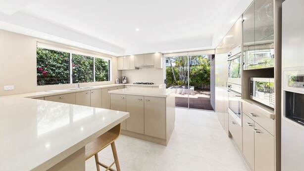 A modern kitchen with a central island, built-in appliances, and a view of the garden. - Luxury Riverfront: Private Pool, Water Views (Noosa Heads)