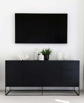 Suporte de TV preto com televisão de tela plana montada acima, um vaso de planta e objetos decorativos.