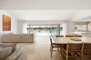 A modern living room with a large window overlooking a lake, a dining area with wooden chairs, and a kitchen with a large island. - The River Rest - Noosa Jetty Stay (Noosa Heads)
