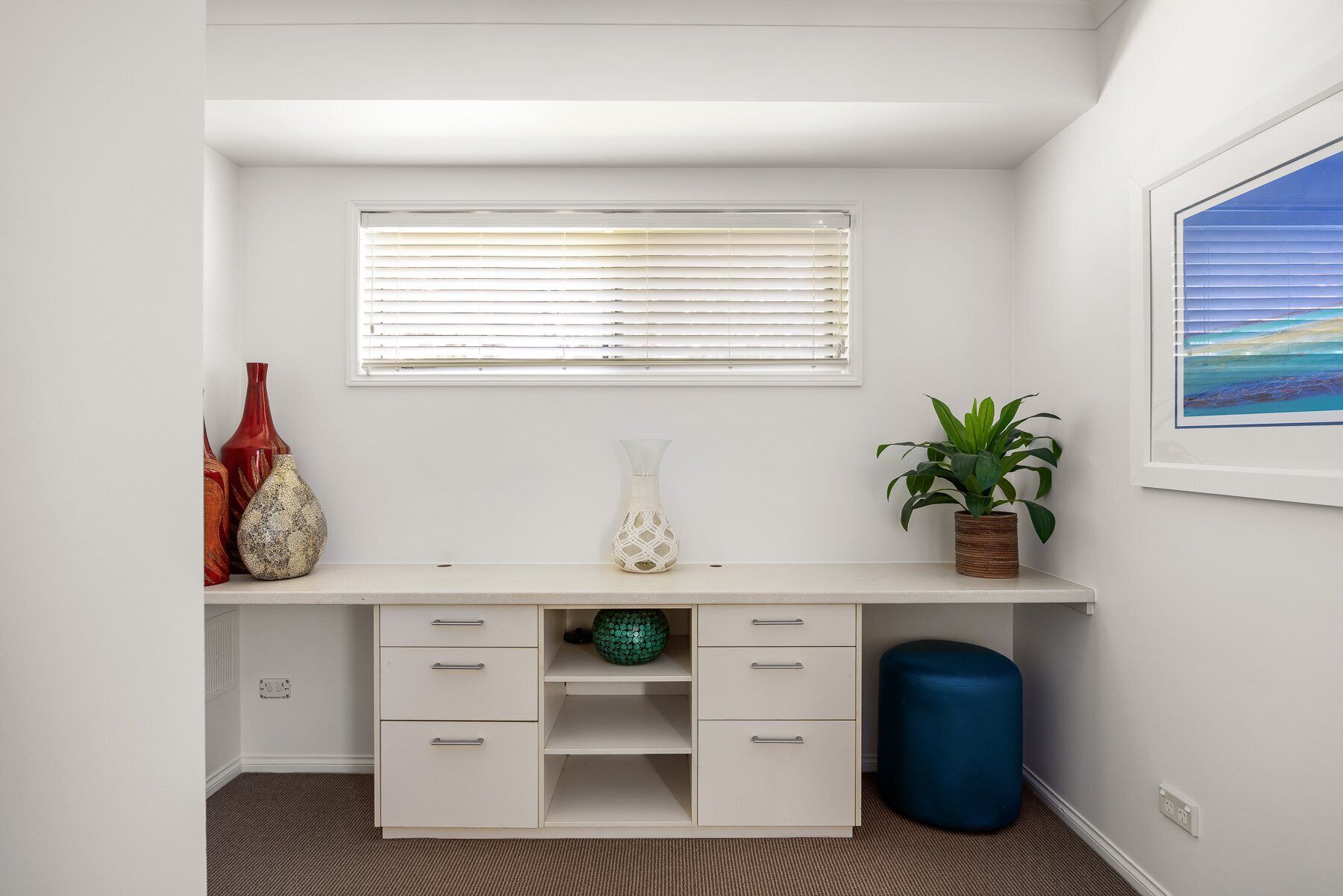 Un bureau blanc avec des tiroirs, une fenêtre avec des stores, une plante verte et une poubelle bleue.