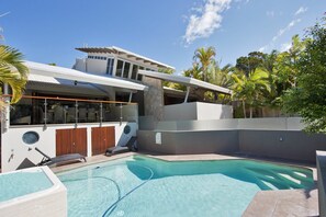 Un área de piscina moderna con una piscina cristalina, sillas de descanso y un espacio de estar cubierto.