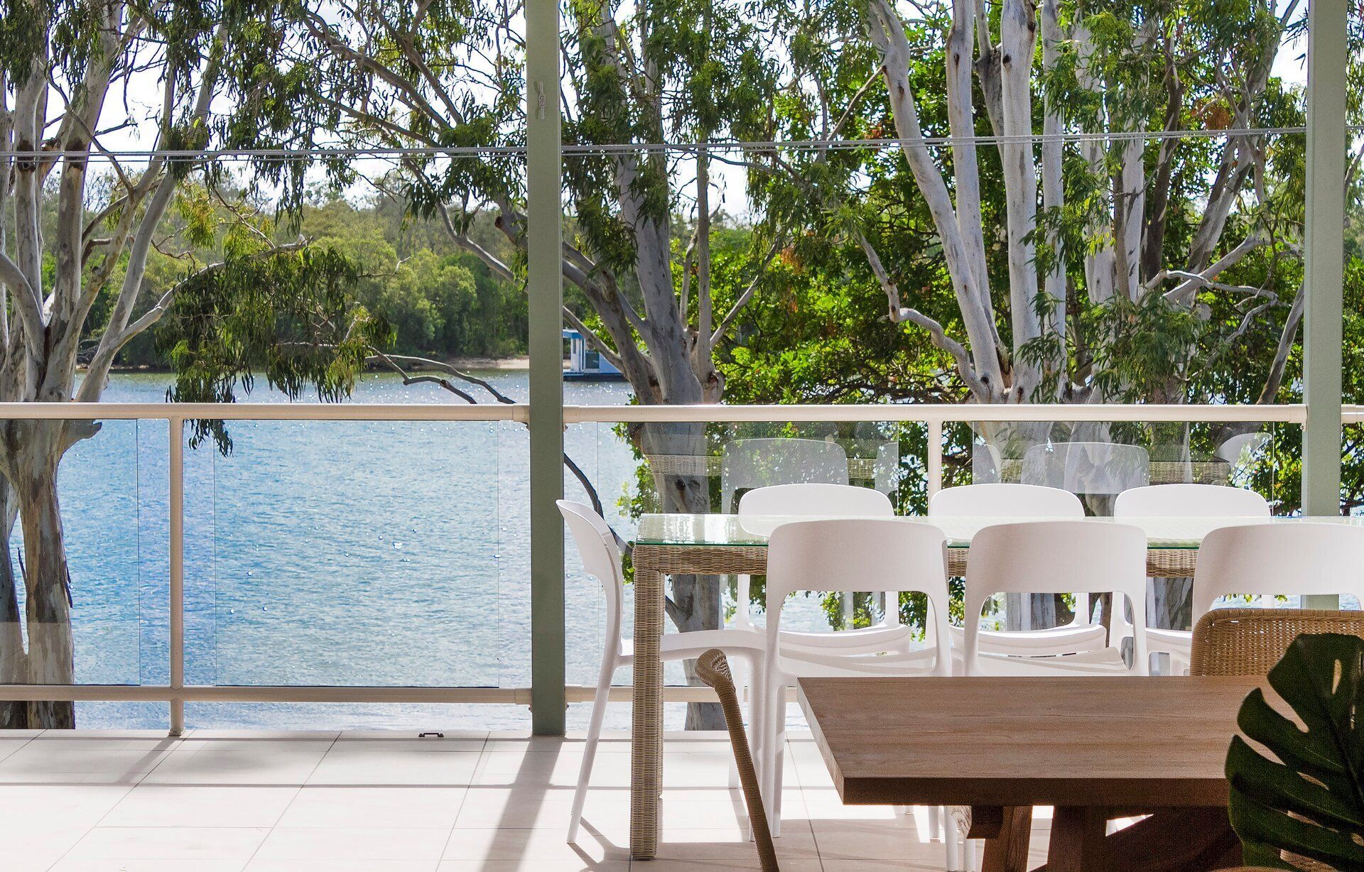 Una terraza con vista a un lago, rodeada de árboles, con sillas de plástico blanco y una mesa de madera.