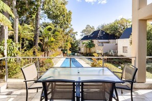 Un área de estar al aire libre con una mesa y sillas de vidrio, con vistas a una piscina y un jardín ajardinado con diversas árboles y arbustos.